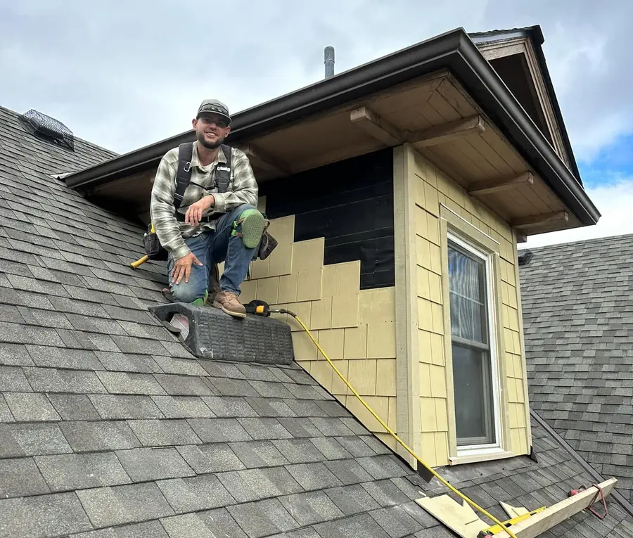 Timberwolf crew member sitting on a freshly shingled roof beside a dormer under repair